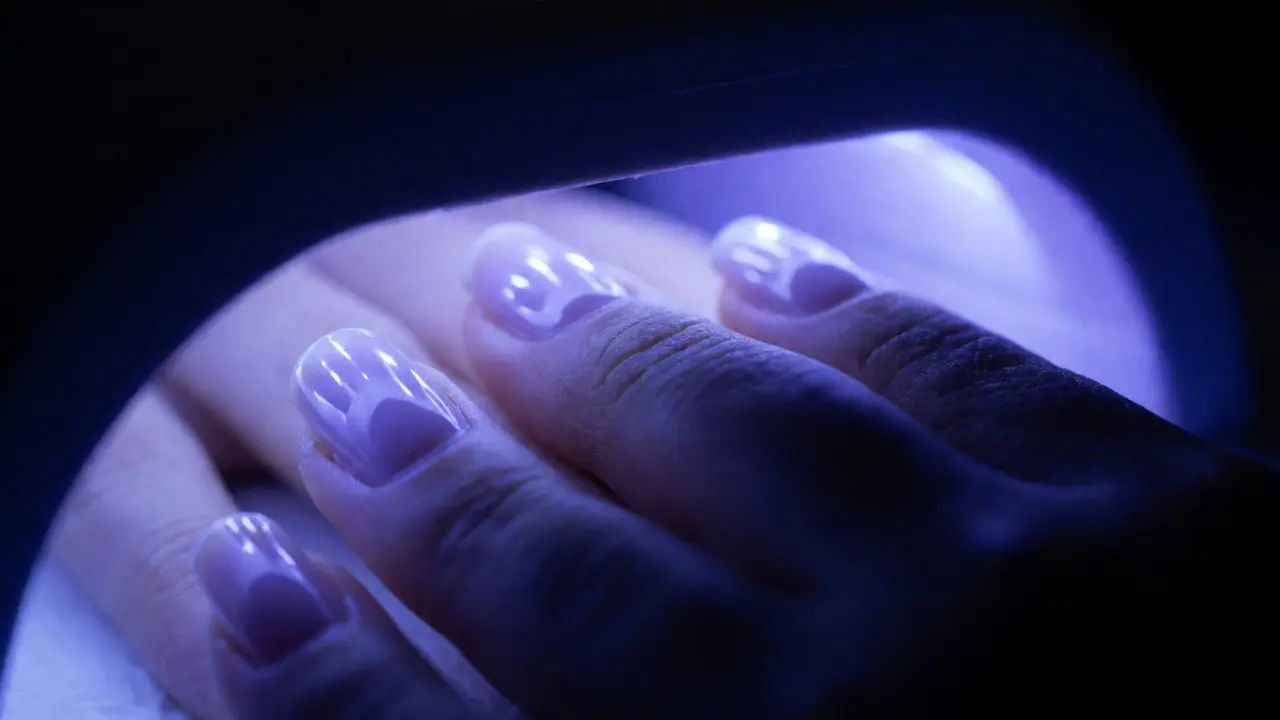Hand under a UV LED lamp curing gel polish with a violet glow.